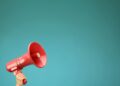 A person's hand holds up a bright red plastic megaphone or bullhorn in the lower left corner against a solid, vibrant teal background. The megaphone is angled upward toward the right, leaving ample negative space in the upper portion of the frame.