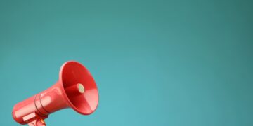 A person's hand holds up a bright red plastic megaphone or bullhorn in the lower left corner against a solid, vibrant teal background. The megaphone is angled upward toward the right, leaving ample negative space in the upper portion of the frame.