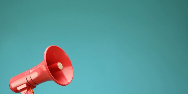 A person's hand holds up a bright red plastic megaphone or bullhorn in the lower left corner against a solid, vibrant teal background. The megaphone is angled upward toward the right, leaving ample negative space in the upper portion of the frame.