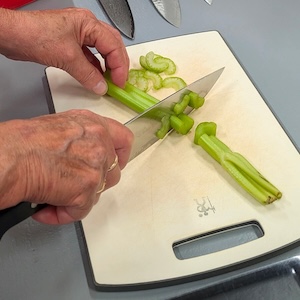 A close-up view of a person's hands using a large chef's knife to chop green celery stalks on a cream-colored cutting board. Several sliced pieces of celery sit clustered at the top of the cutting board, while a couple of unsliced stalks lie ready next to the blade.