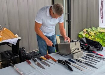 A man in a white polo shirt and jeans leans over an outdoor table, demonstrating an electric knife-sharpening machine. The table is covered in a light blue cloth and laid out with a variety of kitchen knives, chef knives, and sharpening tools. In the background, a small table holds a wooden knife block, a white banner with a logo stands nearby, and green garden plants are visible to the right.