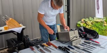 A man in a white polo shirt and jeans leans over an outdoor table, demonstrating an electric knife-sharpening machine. The table is covered in a light blue cloth and laid out with a variety of kitchen knives, chef knives, and sharpening tools. In the background, a small table holds a wooden knife block, a white banner with a logo stands nearby, and green garden plants are visible to the right.
