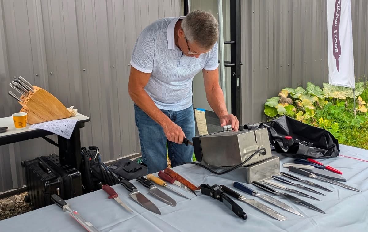 A man in a white polo shirt and jeans leans over an outdoor table, demonstrating an electric knife-sharpening machine. The table is covered in a light blue cloth and laid out with a variety of kitchen knives, chef knives, and sharpening tools. In the background, a small table holds a wooden knife block, a white banner with a logo stands nearby, and green garden plants are visible to the right.