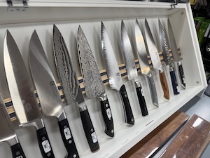 A row of premium culinary kitchen knives organized neatly inside a white display case. The selection features various blades including standard stainless steel chef's knives, distinct textured Damascus steel blades, and carving knives with varied wooden, black, and blue handles.