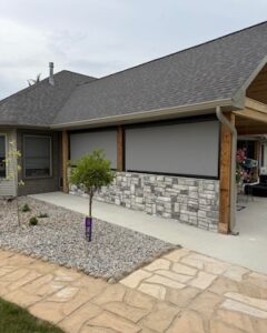 Two large grey motorized retractable shades lowered to enclose a modern covered patio structure attached to a house. The lower portion of the patio wall features a decorative grey faux stone veneer panel. In the foreground, a paved stone walkway runs alongside a graveled landscaping bed containing a small young tree.