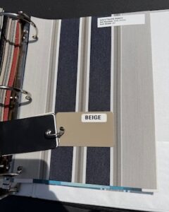 A three-ring binder open to a fabric sample page showing a striped pattern in navy blue, beige, and white. A label in the upper right reads "NAVY/TAUPE ERAD-S". Two smaller color chips are attached to the ring mechanism over the page, including a tan chip labeled "BEIGE" and a dark navy chip.