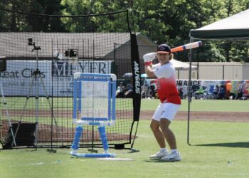 Outdoor action shot of a wiffle ball player preparing for a pitch on a vibrant green turf field. The player wears a blue and red baseball cap with a "T" logo, a white athletic shirt transitioning into a red gradient at the bottom with the number 12, white shorts, and white sneakers. He smiles broadly while holding a blue, white, and orange plastic bat in a classic batting stance. Behind him sits an Aquigelis black backstop net, a blue and white custom PVC pipe strike zone target, and an outfield wall lined with local business sponsor banners including Great Clips and Meijer.