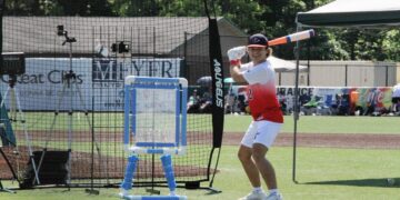 Outdoor action shot of a wiffle ball player preparing for a pitch on a vibrant green turf field. The player wears a blue and red baseball cap with a "T" logo, a white athletic shirt transitioning into a red gradient at the bottom with the number 12, white shorts, and white sneakers. He smiles broadly while holding a blue, white, and orange plastic bat in a classic batting stance. Behind him sits an Aquigelis black backstop net, a blue and white custom PVC pipe strike zone target, and an outfield wall lined with local business sponsor banners including Great Clips and Meijer.