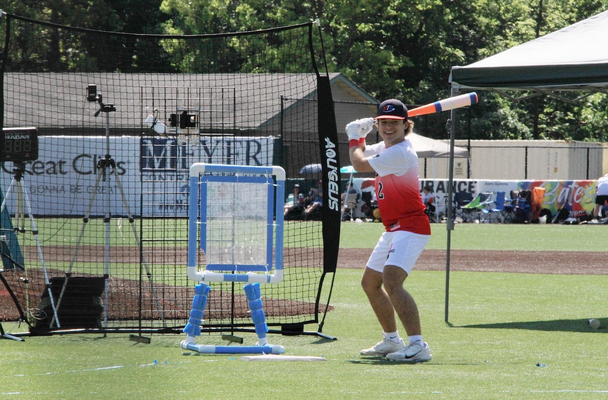 Outdoor action shot of a wiffle ball player preparing for a pitch on a vibrant green turf field. The player wears a blue and red baseball cap with a "T" logo, a white athletic shirt transitioning into a red gradient at the bottom with the number 12, white shorts, and white sneakers. He smiles broadly while holding a blue, white, and orange plastic bat in a classic batting stance. Behind him sits an Aquigelis black backstop net, a blue and white custom PVC pipe strike zone target, and an outfield wall lined with local business sponsor banners including Great Clips and Meijer.