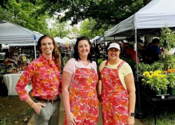 Three smiling people stand outdoors on a paved path lined with white vendor tents at a busy farmers market. On the left, a person with long light brown hair wears a vibrant button-down shirt with a bright pink and orange floral pattern, tucked into grey trousers with a brown belt. In the center, a woman wearing a pink t-shirt poses in denim overalls featuring the identical pink and orange floral print. On the right, a woman wearing a yellow t-shirt and a white visor wears a matching short apron or pinafore in the same floral pattern. Potted yellow marigolds and lush green foliage sit on a table in the background.