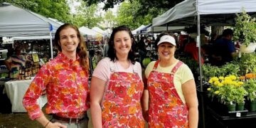Three smiling people stand outdoors on a paved path lined with white vendor tents at a busy farmers market. On the left, a person with long light brown hair wears a vibrant button-down shirt with a bright pink and orange floral pattern, tucked into grey trousers with a brown belt. In the center, a woman wearing a pink t-shirt poses in denim overalls featuring the identical pink and orange floral print. On the right, a woman wearing a yellow t-shirt and a white visor wears a matching short apron or pinafore in the same floral pattern. Potted yellow marigolds and lush green foliage sit on a table in the background.