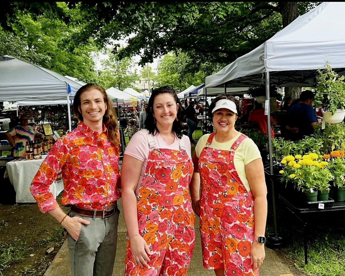 Three smiling people stand outdoors on a paved path lined with white vendor tents at a busy farmers market. On the left, a person with long light brown hair wears a vibrant button-down shirt with a bright pink and orange floral pattern, tucked into grey trousers with a brown belt. In the center, a woman wearing a pink t-shirt poses in denim overalls featuring the identical pink and orange floral print. On the right, a woman wearing a yellow t-shirt and a white visor wears a matching short apron or pinafore in the same floral pattern. Potted yellow marigolds and lush green foliage sit on a table in the background.