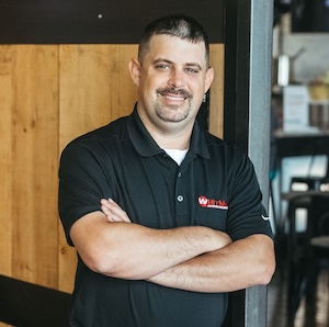 A portrait of a smiling man with a goatee and short dark hair, standing with his arms crossed while leaning against a doorway. He is wearing a black polo shirt with a red and white Thiele Heating & Air Conditioning logo on the left chest, with a light-colored wood paneled wall behind him.