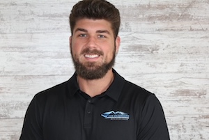 A portrait of a smiling young man with dark brown hair and a thick beard, wearing a black polo shirt featuring a blue and white mountain-style logo on the left chest, posing against a white, rustic wood-plank wall background.