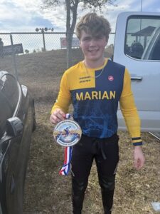 A smiling young male Marian University collegiate BMX racer stands proudly outdoors holding a championship medal. He is wearing a blue and yellow long-sleeve racing jersey with "MARIAN" printed across the chest in bold yellow letters, paired with black pants. He holds the circular medal by its red, white, and blue striped ribbon. A silver pickup truck and a dark sedan are visible in the background.