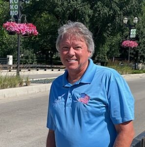 An outdoor portrait of a smiling man with graying hair wearing a blue polo shirt with an embroidered American flag on the left chest, standing on a street in Kokomo, Indiana, with green trees and pink hanging flower baskets in the background.