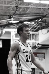 A black-and-white, medium close-up portrait of a young male basketball player looking off to the side during a game. He is wearing a white basketball jersey with dark trim, featuring the cursive text "Daleville" and the number "21" on the front. He is inside a gymnasium with overhead metal scaffolding, rafters, and lights visible in the background.