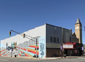 A street-level corner view of the historic Diana Theatre building under a clear blue sky. The front facade features a dark marquee canopy with red lettering and a white reader board. The expansive side wall of the building is decorated with a vibrant, colorful mural depicting local heritage elements, carnival shapes, and a large blue vertical ticket graphic that reads "ADMIT ONE." In the background, the tall brick clock tower of the Tipton County Courthouse rises above the street corner next to a traffic light.