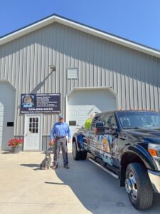 A business owner and a grey dog stand on a concrete driveway in front of a gray corrugated metal commercial building. To the right, a black Ford Super Duty dually service truck displays a custom commercial vehicle wrap. A company sign on the building features service details, a logo, and the phone number 574-290-0240.
