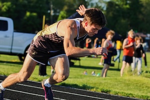 An intense, close-up action shot captures a male track athlete mid-stride as he bursts forward from the starting line. He leans low with explosive power, wearing a dark brown and white running singlet and matching athletic shorts. He is focused intently forward on a black running track. The background shows a grassy field with blurred spectators, trees, and a white pickup truck illuminated by bright, late-afternoon sunlight.