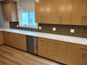 A modern kitchen featuring light oak wood cabinetry with long, silver bar handles. The countertops are clean, solid white quartz, housing an under-mount sink with a chrome faucet. A dark olive green herringbone tile backsplash runs between the upper and lower cabinets. A stainless steel dishwasher is integrated into the lower cabinetry row over light wood floors.