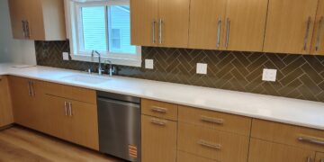 A modern kitchen featuring light oak wood cabinetry with long, silver bar handles. The countertops are clean, solid white quartz, housing an under-mount sink with a chrome faucet. A dark olive green herringbone tile backsplash runs between the upper and lower cabinets. A stainless steel dishwasher is integrated into the lower cabinetry row over light wood floors.