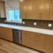A modern kitchen featuring light oak wood cabinetry with long, silver bar handles. The countertops are clean, solid white quartz, housing an under-mount sink with a chrome faucet. A dark olive green herringbone tile backsplash runs between the upper and lower cabinets. A stainless steel dishwasher is integrated into the lower cabinetry row over light wood floors.