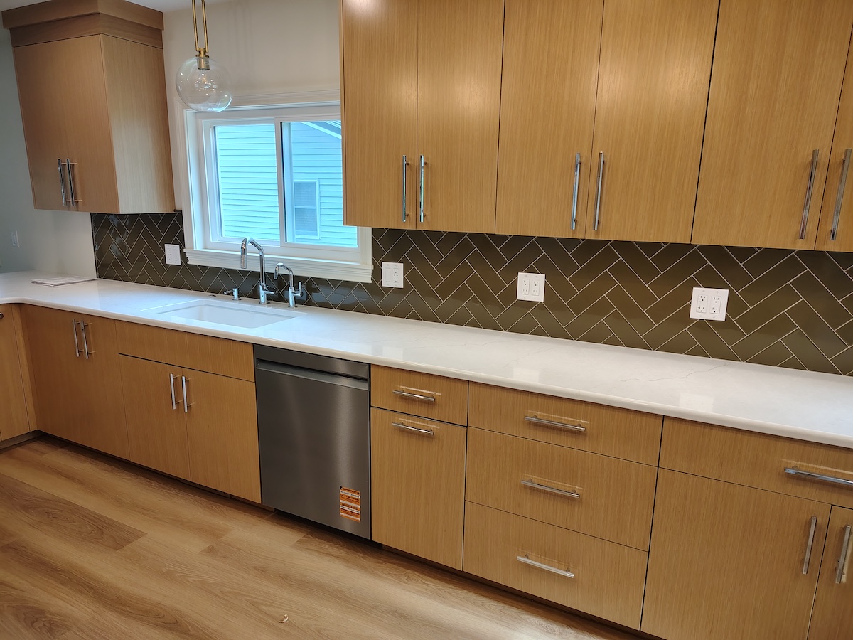 A modern kitchen featuring light oak wood cabinetry with long, silver bar handles. The countertops are clean, solid white quartz, housing an under-mount sink with a chrome faucet. A dark olive green herringbone tile backsplash runs between the upper and lower cabinets. A stainless steel dishwasher is integrated into the lower cabinetry row over light wood floors.
