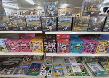 Retail store shelves filled with a variety of jigsaw puzzles and children's activity books. The top shelf displays 1,000-piece landscape puzzles from Heartwood, including themes like barns and lighthouses. The middle shelf holds multi-pack puzzle sets from brands like Tootsie Roll, Pringles, and Ceaco. The bottom shelf features educational activity items, sticker books, and dry-erase pockets.