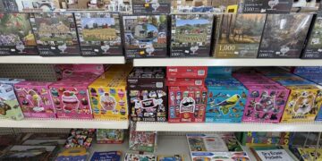 Retail store shelves filled with a variety of jigsaw puzzles and children's activity books. The top shelf displays 1,000-piece landscape puzzles from Heartwood, including themes like barns and lighthouses. The middle shelf holds multi-pack puzzle sets from brands like Tootsie Roll, Pringles, and Ceaco. The bottom shelf features educational activity items, sticker books, and dry-erase pockets.