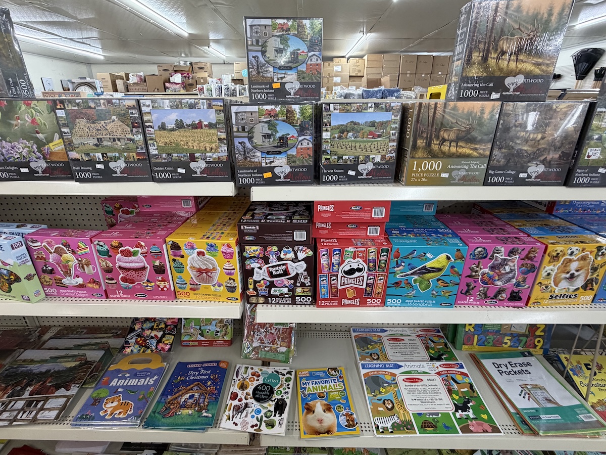 Retail store shelves filled with a variety of jigsaw puzzles and children's activity books. The top shelf displays 1,000-piece landscape puzzles from Heartwood, including themes like barns and lighthouses. The middle shelf holds multi-pack puzzle sets from brands like Tootsie Roll, Pringles, and Ceaco. The bottom shelf features educational activity items, sticker books, and dry-erase pockets.