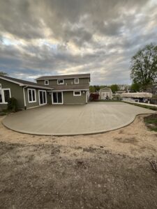 A large, newly poured concrete patio with smooth, sweeping curved edges in the backyard of a two-story olive green house. The wide concrete pad fills the outdoor space between the home, a backyard storage shed, and a boat parked on a trailer. A dramatic, cloudy evening sky hangs overhead.