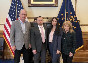 Full-length indoor photograph of four individuals standing shoulder-to-shoulder inside an executive office. From left to right: Indiana Governor Mike Braun wears a light gray blazer over a light blue button-down shirt and black pants; a man wears a dark gray suit with a light pink tie; a woman wears a navy blue pant suit over a pink-and-white striped blouse; and a woman wears a dark green-and-navy plaid blazer with black pants. They stand on a blue carpet with a yellow geometric border pattern in front of a marble fireplace mantel. Flanking the group are the American flag on the left and the dark blue Indiana state flag on the right. Centered on the wall above the fireplace is a large, ornate gold frame holding an oil portrait of a historical bearded man in a formal suit.