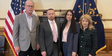 Full-length indoor photograph of four individuals standing shoulder-to-shoulder inside an executive office. From left to right: Indiana Governor Mike Braun wears a light gray blazer over a light blue button-down shirt and black pants; a man wears a dark gray suit with a light pink tie; a woman wears a navy blue pant suit over a pink-and-white striped blouse; and a woman wears a dark green-and-navy plaid blazer with black pants. They stand on a blue carpet with a yellow geometric border pattern in front of a marble fireplace mantel. Flanking the group are the American flag on the left and the dark blue Indiana state flag on the right. Centered on the wall above the fireplace is a large, ornate gold frame holding an oil portrait of a historical bearded man in a formal suit.