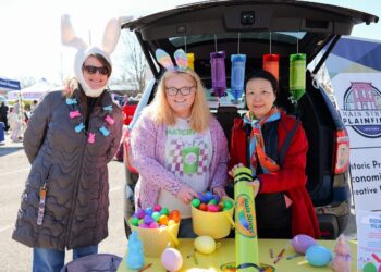 Three women smile beside the open trunk of a black SUV during a festive outdoor community event. On the left, a woman in a long brown puffer jacket wears a white bunny ears hat and a pastel rabbit necklace. In the center, a woman with blonde hair and glasses wears a pink cardigan, a graphic t-shirt, and tie-dye bunny ears, holding a yellow basket filled with colorful plastic Easter eggs. On the right, a woman in a red jacket and an orange scarf holds a large lime green toy crayon labeled "Main Street Plainfield." Large decorative crayons hang from the trunk liftgate above a table featuring more decorative eggs.