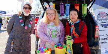 Three women smile beside the open trunk of a black SUV during a festive outdoor community event. On the left, a woman in a long brown puffer jacket wears a white bunny ears hat and a pastel rabbit necklace. In the center, a woman with blonde hair and glasses wears a pink cardigan, a graphic t-shirt, and tie-dye bunny ears, holding a yellow basket filled with colorful plastic Easter eggs. On the right, a woman in a red jacket and an orange scarf holds a large lime green toy crayon labeled "Main Street Plainfield." Large decorative crayons hang from the trunk liftgate above a table featuring more decorative eggs.