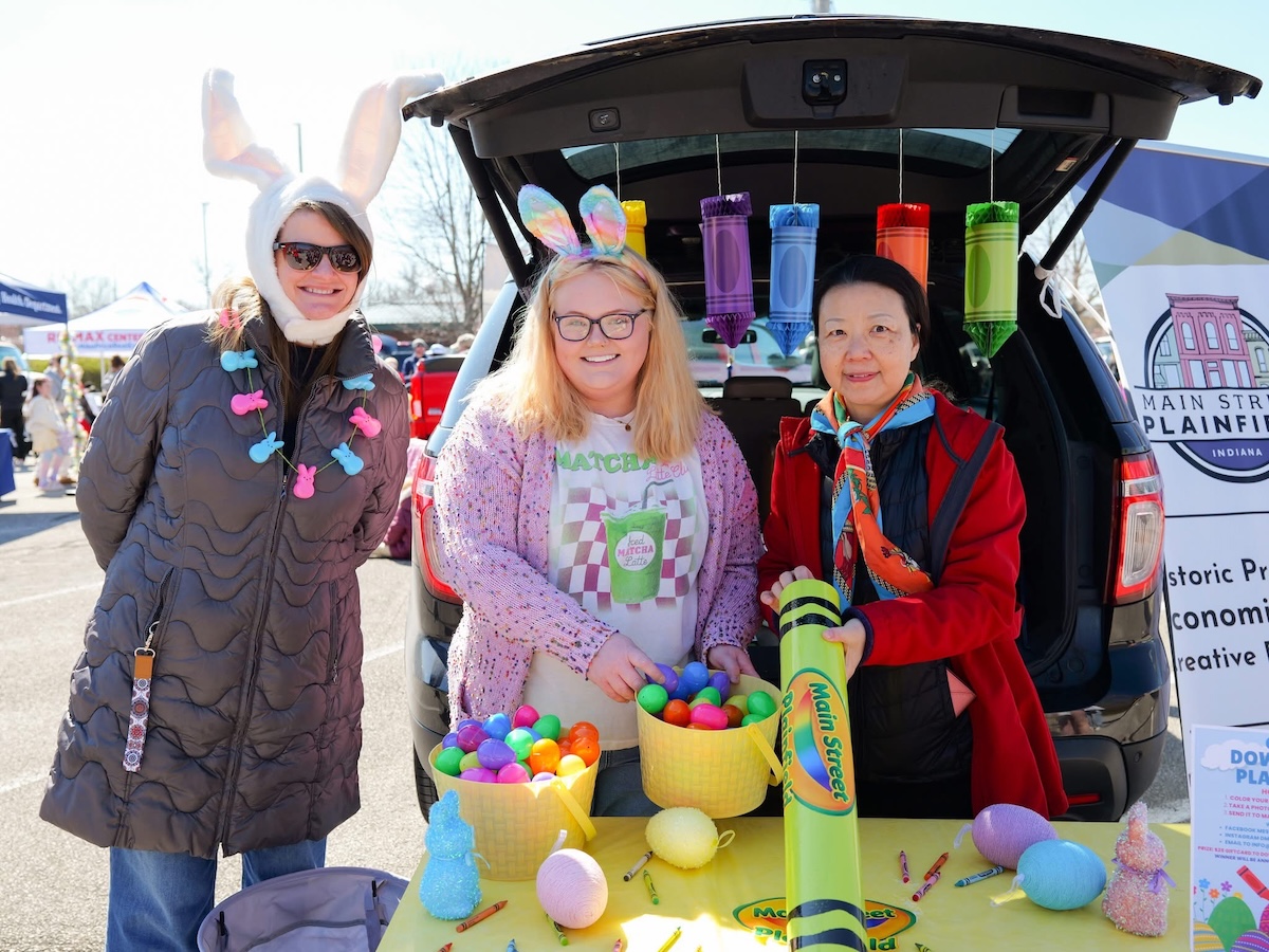 Three women smile beside the open trunk of a black SUV during a festive outdoor community event. On the left, a woman in a long brown puffer jacket wears a white bunny ears hat and a pastel rabbit necklace. In the center, a woman with blonde hair and glasses wears a pink cardigan, a graphic t-shirt, and tie-dye bunny ears, holding a yellow basket filled with colorful plastic Easter eggs. On the right, a woman in a red jacket and an orange scarf holds a large lime green toy crayon labeled "Main Street Plainfield." Large decorative crayons hang from the trunk liftgate above a table featuring more decorative eggs.