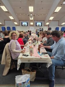 A large group of people sits at long tables inside a crowded event hall playing a game. In the foreground, a woman with curly blonde hair and glasses smiles towards the camera from her seat at a white-clothed table decorated with pink accents. Across from her, a man in a light blue button-down shirt and jeans interacts with his table game sheet. The tables stretch deep into the hall beneath a white, angled ceiling with recessed lighting. In the background, two electronic screens display the number "69" above a central stage.