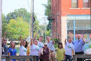 A large group of smiling people stands outdoors on a brick patio, cheering and waving their hands in the air. Most of the group wears matching light blue graphic t-shirts. On the right, a man in a blue checkered button-down shirt waves to the camera. Behind them stands a multi-story red brick building with a large glass storefront window displaying the name "Flowered Occasions." Green trees and power lines are visible under a bright sky in the background.
