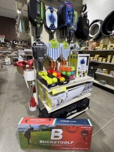 A retail store display stand featuring outdoor recreational sports equipment. The top pegs hang various pickleball paddles, while bins on the shelf below hold bright yellow pickleball balls and multi-colored game balls. A boxed "Official Size Net" sits on the lower shelf. On the floor in front of the display, a large retail box for a "Bucket Golf Bundle" 9-hole set is prominently shown.