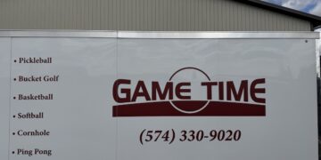 The white exterior side panel of a commercial trailer featuring a large maroon logo that reads "GAME TIME" next to a phone number, (574) 330-9020. A bulleted list on the left side details recreational activities including Pickleball, Bucket Golf, Basketball, Softball, Cornhole, Ping Pong, Volley Ball, and Outdoor Games. A light tan metal barn roof is visible directly behind the top edge of the trailer under a cloudy sky.
