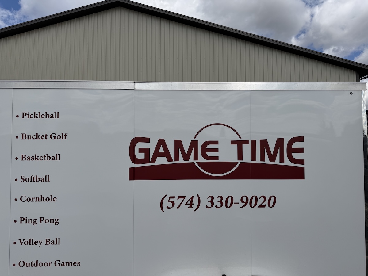 The white exterior side panel of a commercial trailer featuring a large maroon logo that reads "GAME TIME" next to a phone number, (574) 330-9020. A bulleted list on the left side details recreational activities including Pickleball, Bucket Golf, Basketball, Softball, Cornhole, Ping Pong, Volley Ball, and Outdoor Games. A light tan metal barn roof is visible directly behind the top edge of the trailer under a cloudy sky.