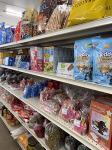 Retail store shelves packed with snacks, cookies, and bagged candies. The top shelves display large bags of chips and pretzels, while the middle shelves hold Teddy Grahams, cookies, and boxes of sweet treats. The lower shelves are filled with clear plastic bags of colorful bulk candies, lollipops, and various pantry items.