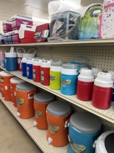 Retail store shelves stocked with various insulated coolers and water jugs. The top shelf displays small handheld hardside coolers and soft lunch bags. The middle shelf is lined with medium-sized insulated personal water jugs in red, blue, lime green, and white. The bottom shelf features large orange, teal, and blue cylindrical water coolers with built-in dispensing spigots.