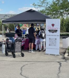 Outdoor shot of a community pop-up event on an asphalt surface under a partly cloudy sky. A black four-legged pop-up canopy tent covers a table draped in a pink and maroon tablecloth, where several people are interacting. A gray utility wagon stroller sits parked on the far left. On the right side of the tent stands a large, white vertical pop-up banner featuring the pink and black logo for "Hailey's Army for Children, Inc." with the text "An Army For Every Child." A large black-and-white QR code labeled "SCAN TO DONATE" sits prominently in the center of the banner above the organization's official website address.