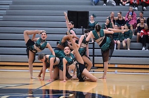 A high school dance team performs a highly synchronized contemporary routine on a polished wooden gymnasium floor. The dancers are grouped tightly together in a dynamic formation, showcasing advanced flexibility with high leg extensions, scorpion extensions, and floor poses. They wear matching dark emerald green and black costumes with sheer mesh details. Spectators sit on grey bleachers in the background watching the performance.