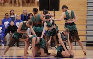 A group of competitive dancers wearing matching emerald green and black costumes perform a contemporary floor routine on a wooden gymnasium court. The dancers are arranged in a tight, layered formation, with some kneeling on the floor, one leaning forward with an outstretched leg, and others standing while interacting through precise choreography. Spectators wearing blue shirts watch from the bleachers in the background next to large audio speakers.