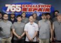 A group photo of twelve employees standing in front of a blue wall inside an office. A large, prominent oval sign behind them reads "765 Collision Repair" in red and white lettering. The team consists of eleven men and one woman, mostly wearing grey sleeveless shirts or t-shirts with a small logo, while a few wear polo shirts or hats. They are lined up side-by-side, smiling directly at the camera.