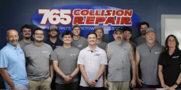 A group photo of twelve employees standing in front of a blue wall inside an office. A large, prominent oval sign behind them reads "765 Collision Repair" in red and white lettering. The team consists of eleven men and one woman, mostly wearing grey sleeveless shirts or t-shirts with a small logo, while a few wear polo shirts or hats. They are lined up side-by-side, smiling directly at the camera.