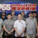 A group photo of twelve employees standing in front of a blue wall inside an office. A large, prominent oval sign behind them reads "765 Collision Repair" in red and white lettering. The team consists of eleven men and one woman, mostly wearing grey sleeveless shirts or t-shirts with a small logo, while a few wear polo shirts or hats. They are lined up side-by-side, smiling directly at the camera.